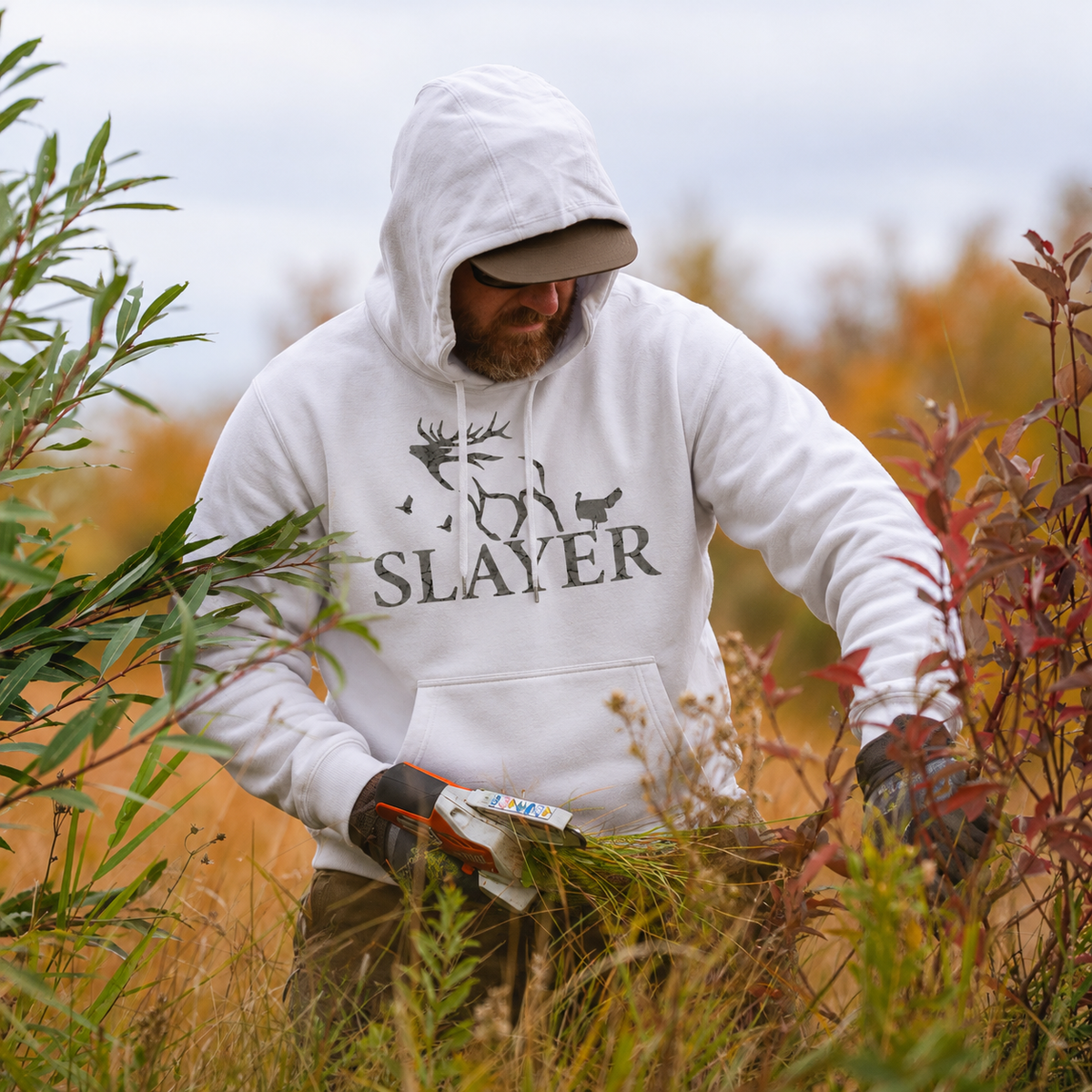 Snow goose hunter wearing a white Slayer Snowbound hoodie while gathering brush among autumn grasses and shrubs.