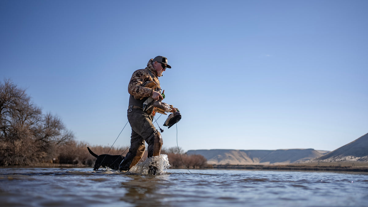 Man in camouflage wading through water with a dog, holding a duck