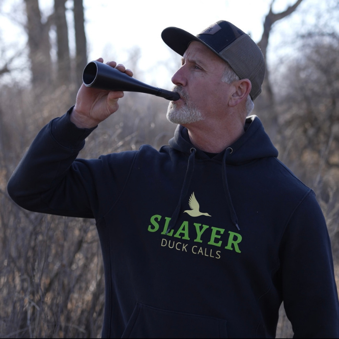 Hunter blowing the Gobbler Slayer turkey call from Slayer Calls, shown in early season woods with branded hoodie.