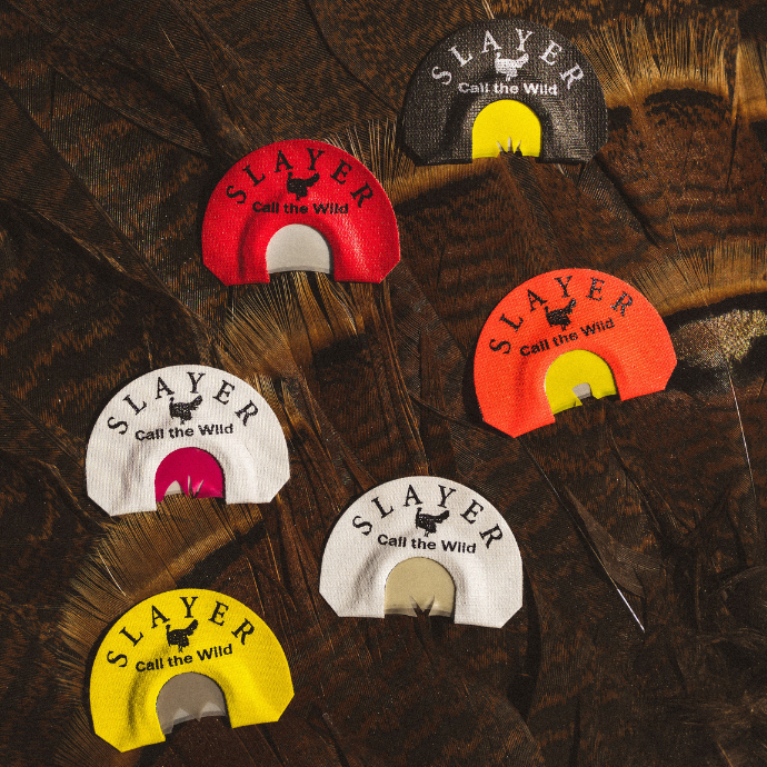 Collection of Slayer turkey mouth calls in various colors and cuts, displayed on a bed of dark turkey feathers — includes batwing, combo cut, and center cut designs.