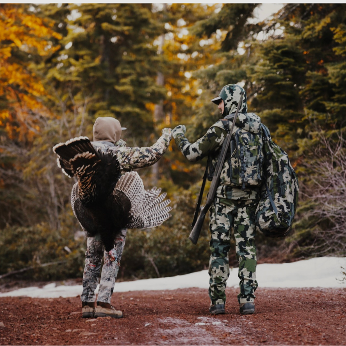 Two camo-clad turkey hunters walking through the woods, one carrying a harvested gobbler, sharing a celebratory fist bump during a successful spring hunt.