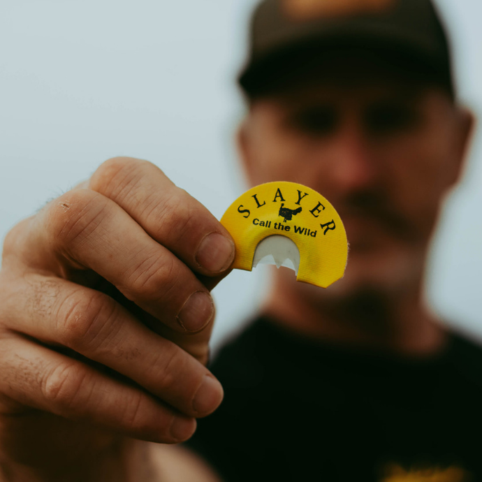 Hunter holding up a Slayer Black Batwing diaphragm turkey call in focus, with a blurred background showcasing the product's detail and branding.