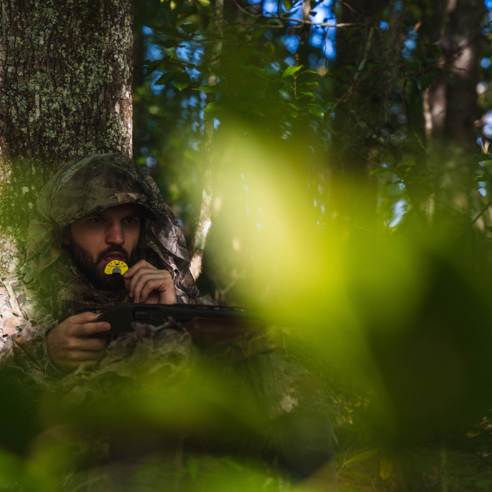 Camouflaged hunter concealed in thick brush, holding a shotgun and using a yellow Slayer Black Batwing diaphragm turkey call during a spring woods setup.