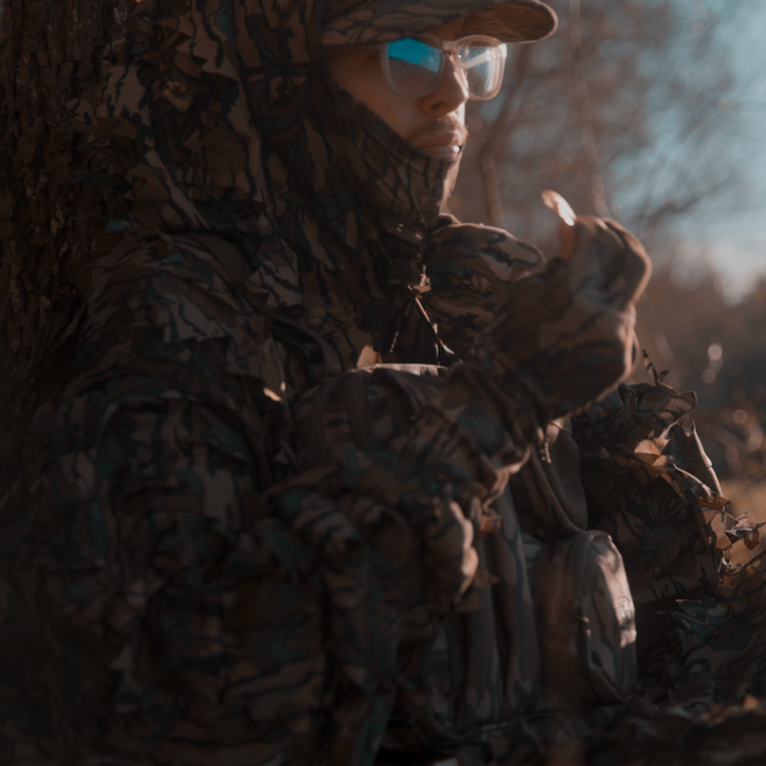 Turkey hunter in full camo gear and glasses sitting against a tree at sunrise, preparing a mouth call while staying concealed in natural cover.