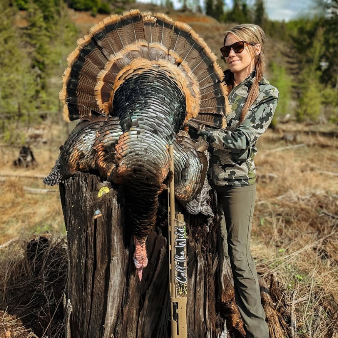 Female hunter in camo standing proudly beside a harvested wild turkey displayed on a tree stump