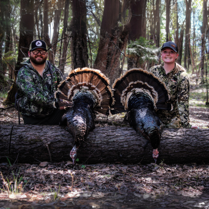 Two hunters in camo pose in the woods with two harvested tom turkeys, fanned out on a fallen log.