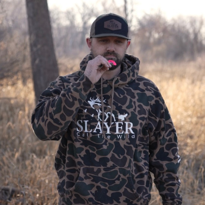 Hunter in camo hoodie using Slayer's Quad Hen turkey mouth call in the field, wearing a Slayer Calls hat.