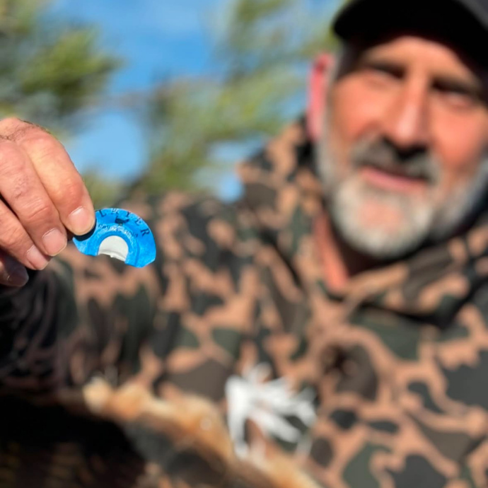 Hunter in camo hoodie holding up Slayer’s The Cutter turkey mouth call in blue, showcasing it proudly in the field on a sunny day.