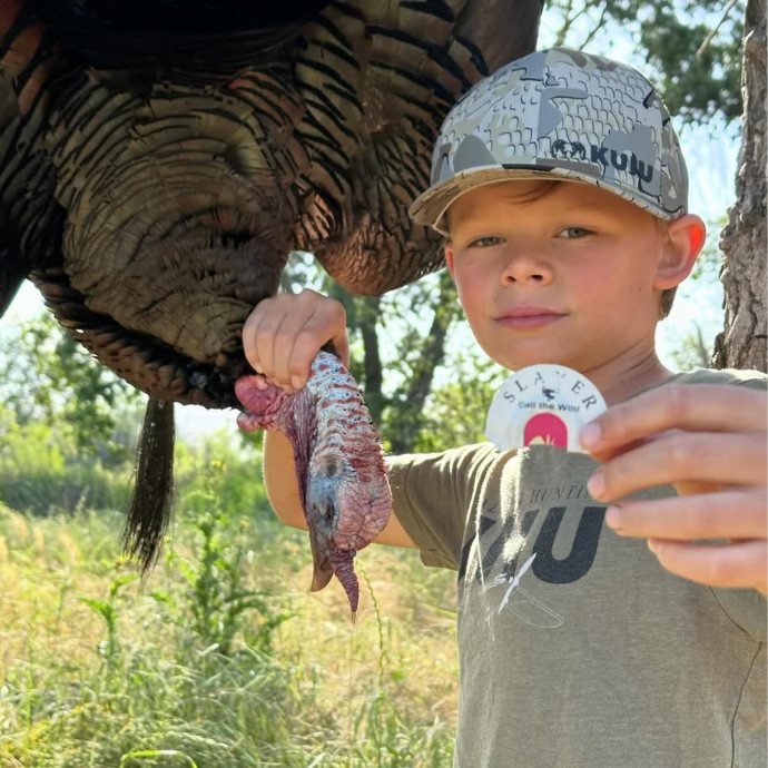 Young hunter proudly holding a harvested wild turkey and a Slayer Fang Cutter mouth call, wearing a KUIU camo hat and standing in the field.