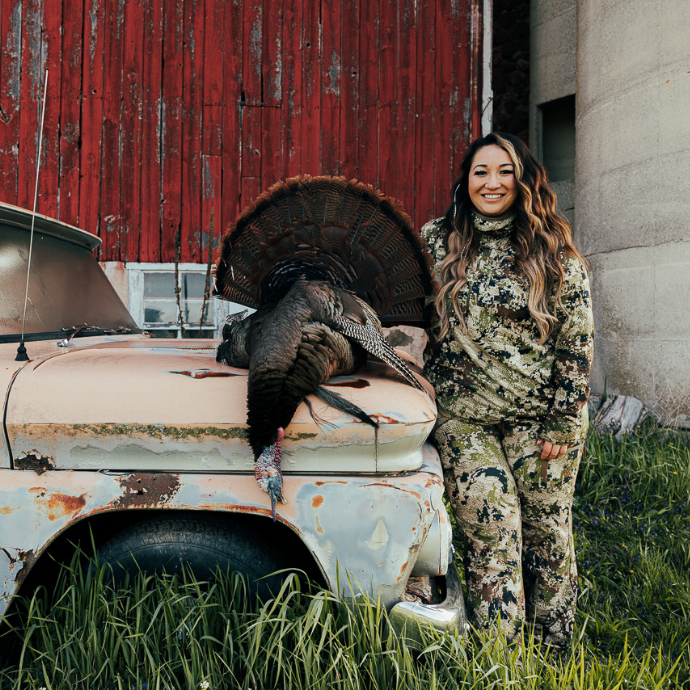 Female hunter in camo poses beside a harvested wild turkey displayed on an old rusted truck, with a red barn and silo in the background.
