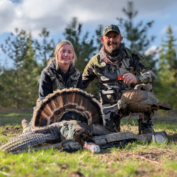Father and daughter pose after a successful turkey hunt, kneeling behind a harvested gobbler with a decoy and Slayer Straight Double diaphragm call in hand.