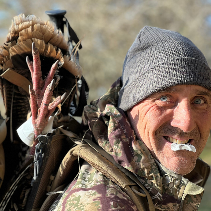 Smiling hunter with a harvested turkey slung over his shoulder and a Slayer Split Triple diaphragm call in his mouth, wearing camo and a beanie in bright morning light.