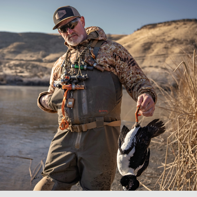 Hunter wearing camo and waders, holding a harvested duck by the feet near a frozen riverbank, with multiple Slayer duck calls on a lanyard across his chest.