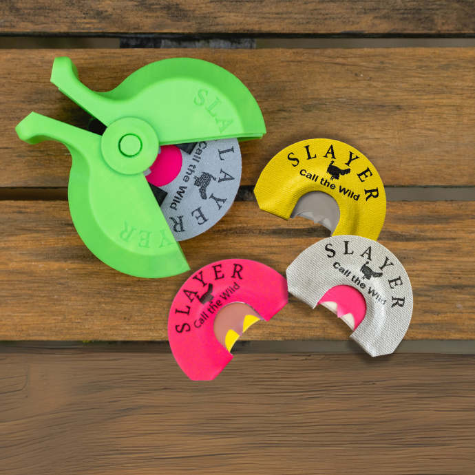 Three Slayer turkey mouth reeds on a wooden surface shown with a bright green Slayer mouth reed holder.