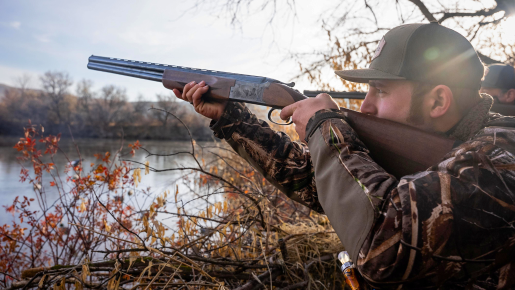 A waterfowl hunter in camouflage is aiming at a flock of mallards coming towards a body of water.
