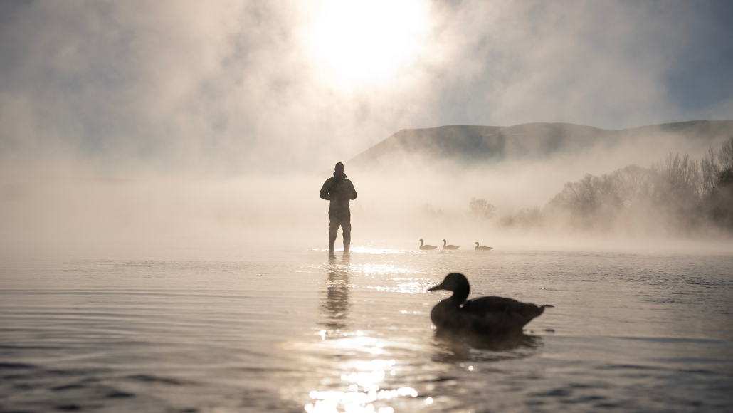 Header image for article featuring best gifts for waterfowlers, featured is a duck hunter wading in the water with decoy sitting in the foreground. Sun rise coming in with fog lifting.