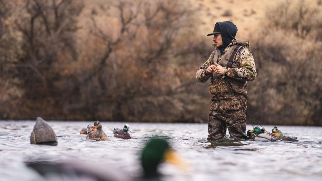 a waterfowl hunter dressed in camo standing in a river putting our duck decoys.