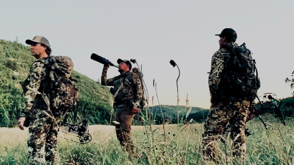 Chance Robbins from God's Country for our Heroes Charity blowing a Slayer bugle tube with 2 other archery elk hunters standing in a field 