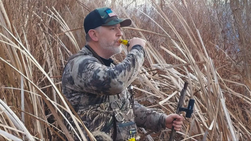 Waterfowl hunter, Travis Tweet is standing in tall grass , blowing a yellow Drake Slayer duck call by Slayer Calls.