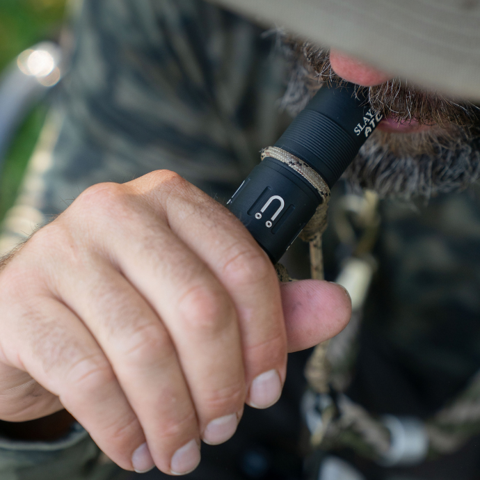 A close-up shot of duck hunter blowing  a black and white single reed ATC - Muller Chokes collaboration duck call with the Muller logo and H2O branding wearing a camouflage shirt in the field.