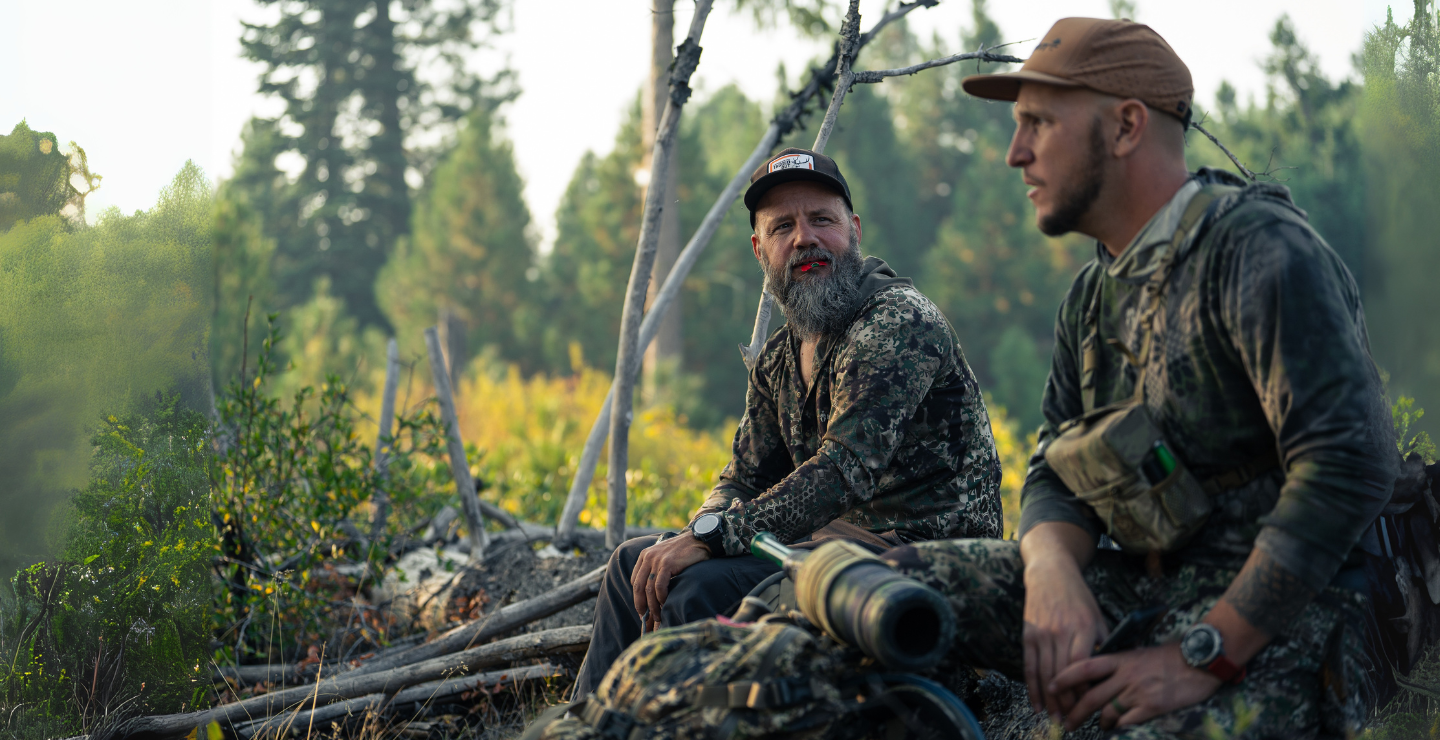 Two hunters in camouflage sitting on a log in a forest setting. One hunter has the Isabella elk reed from Slayer calls in his mouth while the other hunter has the Archangle elk bugle tube sitting in front of him on his pack.