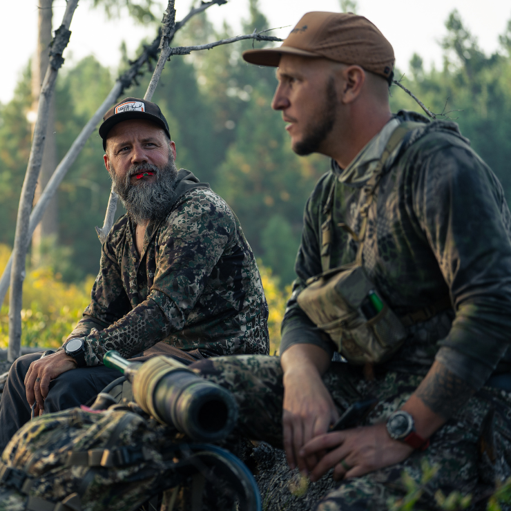 Two hunters in camouflage sitting on a log in a forest setting. One hunter has the Isabella elk reed from Slayer calls in his mouth while the other hunter has the Archangle elk bugle tube sitting in front of him on his pack.
