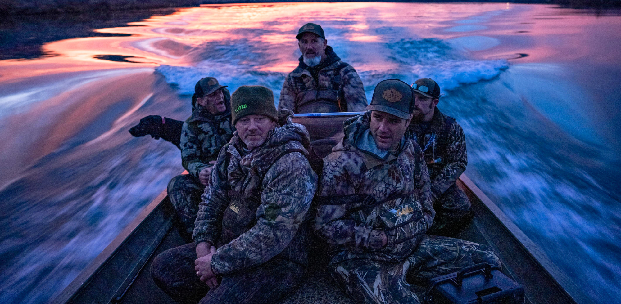 Waterfowl hunters in camo gear and Slayer Calls apparel riding a boat at sunrise on a duck hunting trip.