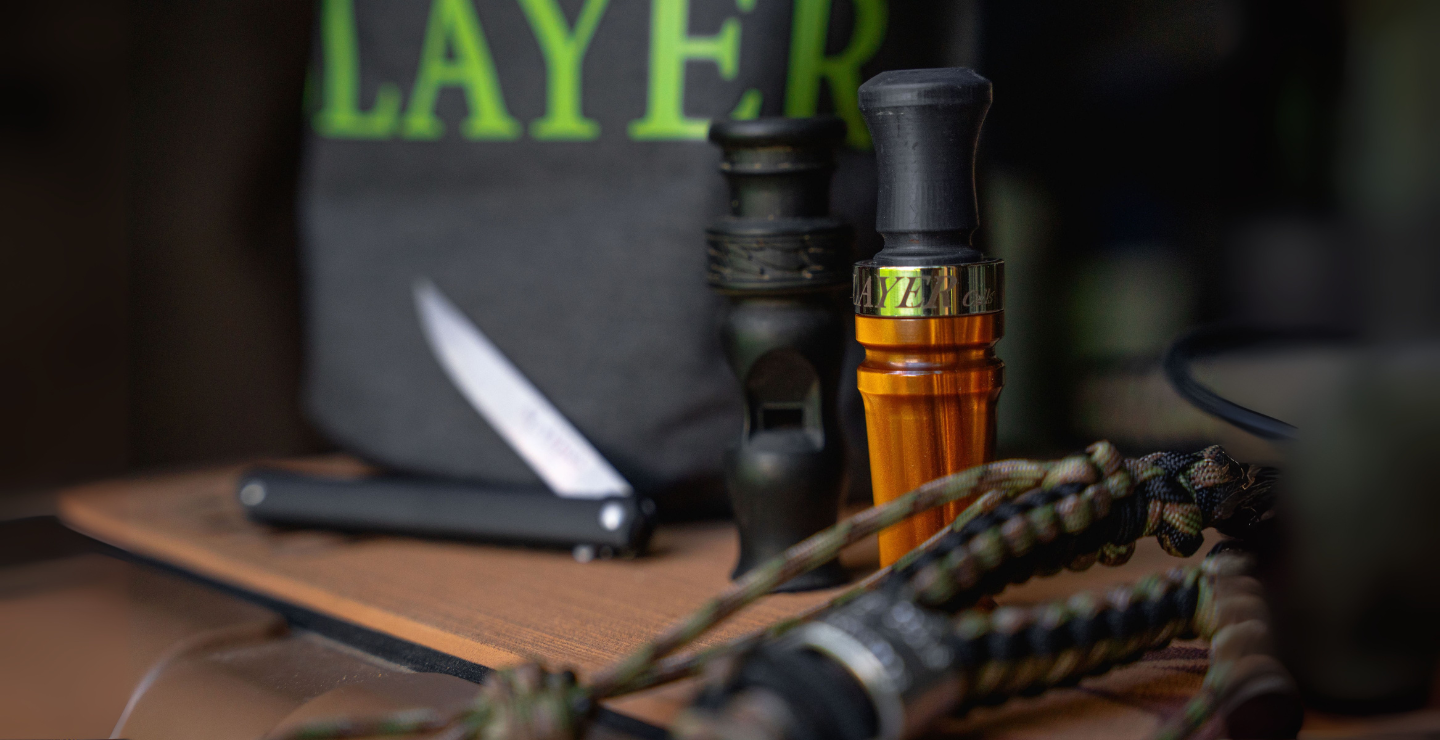 A dark gray Slayer dry bag with a bright green logo and elk silhouette, sitting atop a tan YETI cooler. In front of the bag are two duck calls, a field dresser, and a braided waterfowl lanyard.