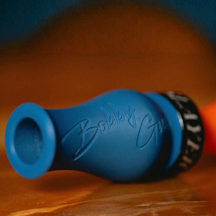 A close-up of the blue and orange Bobby Guy Signature Series duck call with an embossed script signature, a black band reading 'SLAYER' in white, resting on a wooden surface.