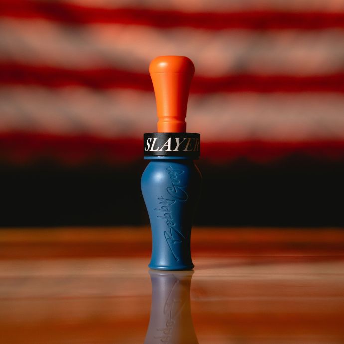 A blue and orange Bobby Guy Signature duck call with black band printed with the word 'SLAYER' stands upright on a wooden surface, with a blurred American flag in the background.