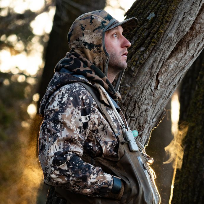 A man in a woodland camouflage hunting jacket and hood stands beside a large tree trunk in a forest, wearing waders and a backpack.