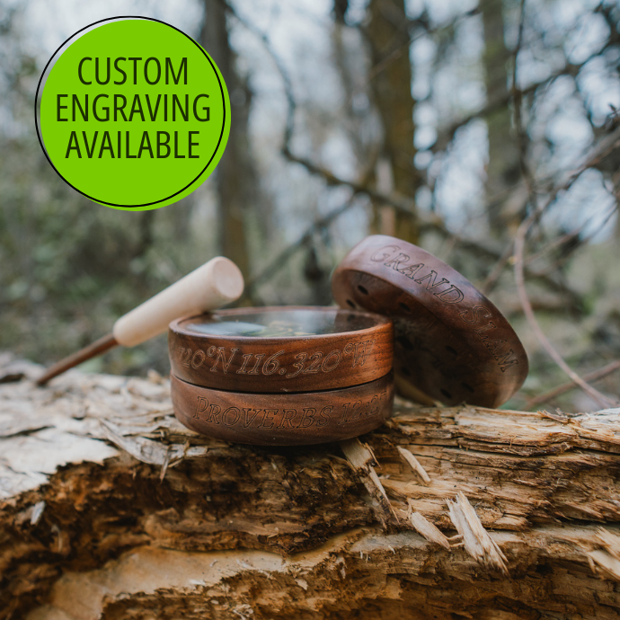 Several wood turkey pod calls with custom engraved coordinates and "Grand Slam" engraving shown on a tree branch in a forest setting, with 'Custom Engraving Available' text.