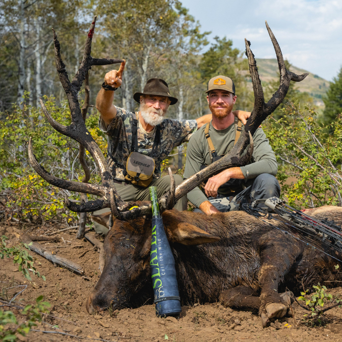 Father and son elk hunters kneeling with mature bull elk called in using Slayer's Logger elk reed from the Clearwater Series and ArchAngel bugle call—successful elk hunting with premium elk calls in the Idaho mountains.