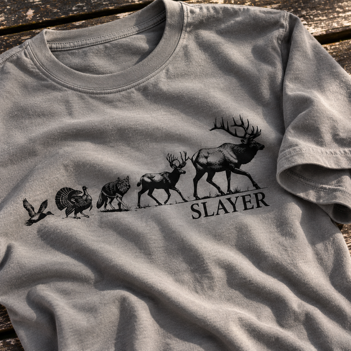 A light gray crewneck t-shirt laid flat on a rustic wooden surface, featuring a black graphic of elk, deer, coyote, turkey and duck silhouettes with the word 'SLAYER' printed underneath.