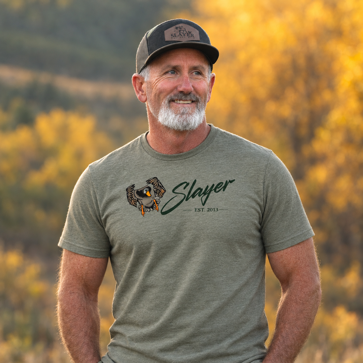 Slayer founder, Bill Ayer wearing an olive green Slayer Heritage T-shirt and Slayer cap outdoors against softly blurred golden autumn foliage.
