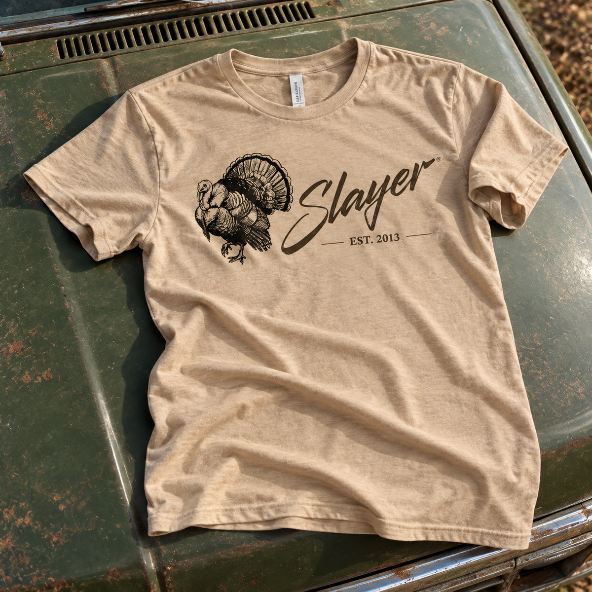 Tan Slayer Longbeard Legacy T-shirt laid on a weathered olive truck hood, showing its turkey graphic.