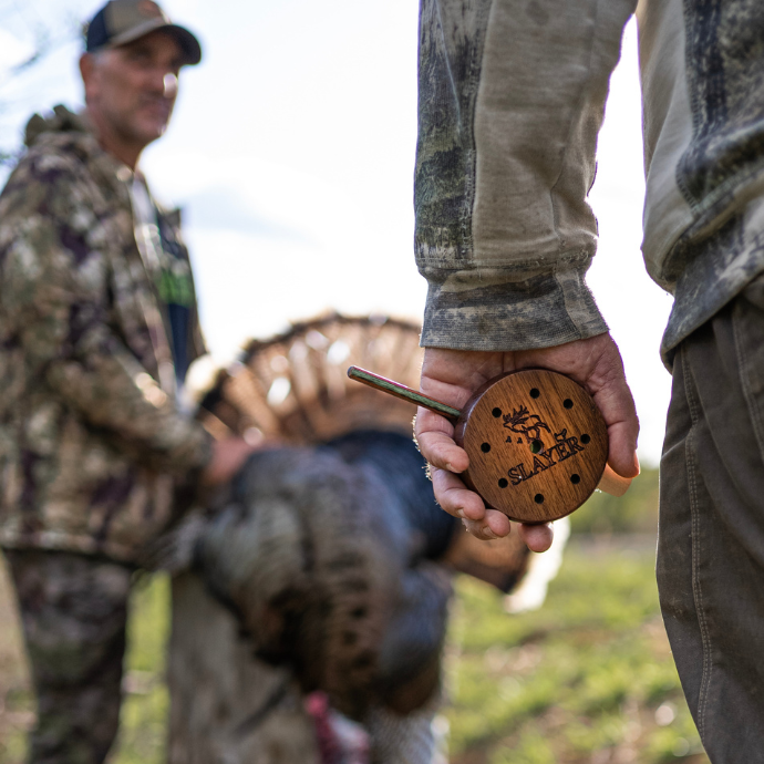 Turkey hunter  holding a wooden pot call with 'Slayer'  branding, with another person in camouflage in the background.