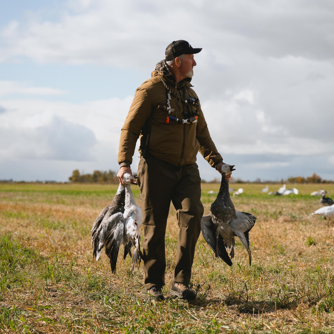 Waterfowl hunter holding several harvested snow geese in a field with a cloudy sky